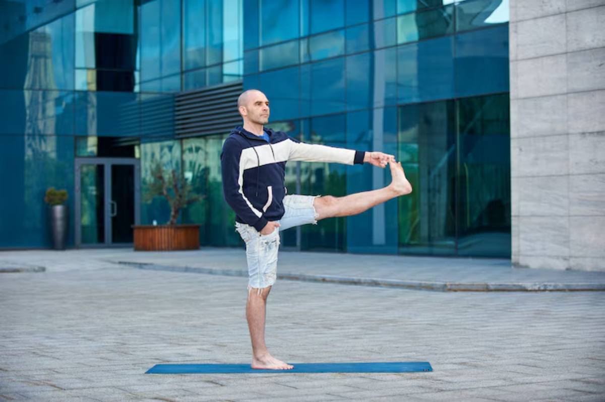 A person practices yoga outdoors, balancing on one foot, holding the other foot with one hand, on a blue mat in a modern urban setting.