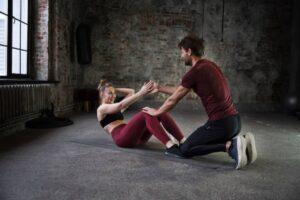 A man assists a woman in a workout session, actively guiding her during a sit-up exercise in a gym setting.