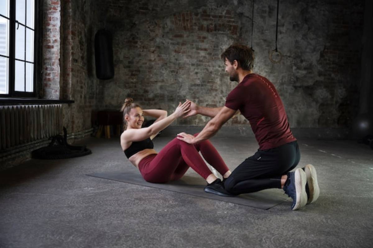 A man assists a woman in a workout session, actively guiding her during a sit-up exercise in a gym setting.