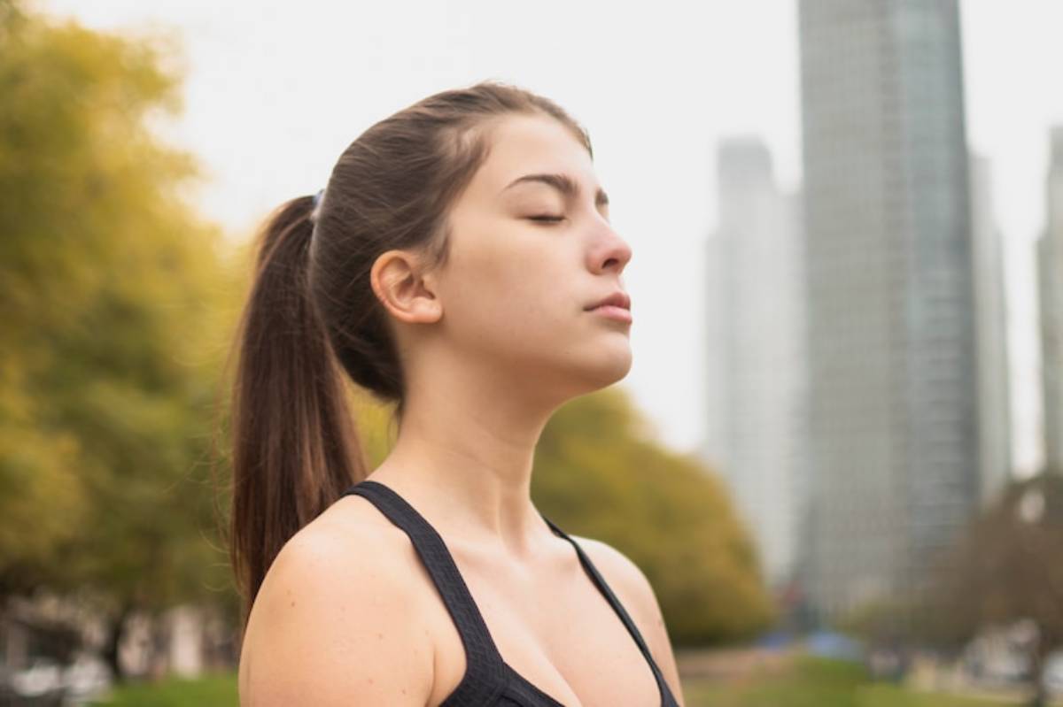 A woman in a black athletic top stands outdoors, gazing upward against a backdrop of city skyscrapers and autumn foliage.
