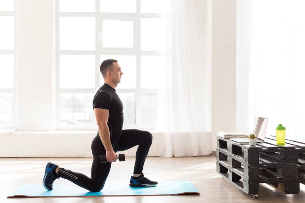 A person performs a lunge on a blue exercise mat, holding a dumbbell, with a laptop and water bottle on a wooden table nearby.