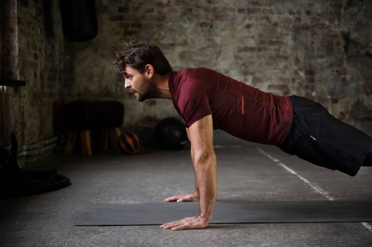 A person in athletic wear performs a push-up on a mat in a rustic gym with exposed brick walls and fitness equipment in the background.