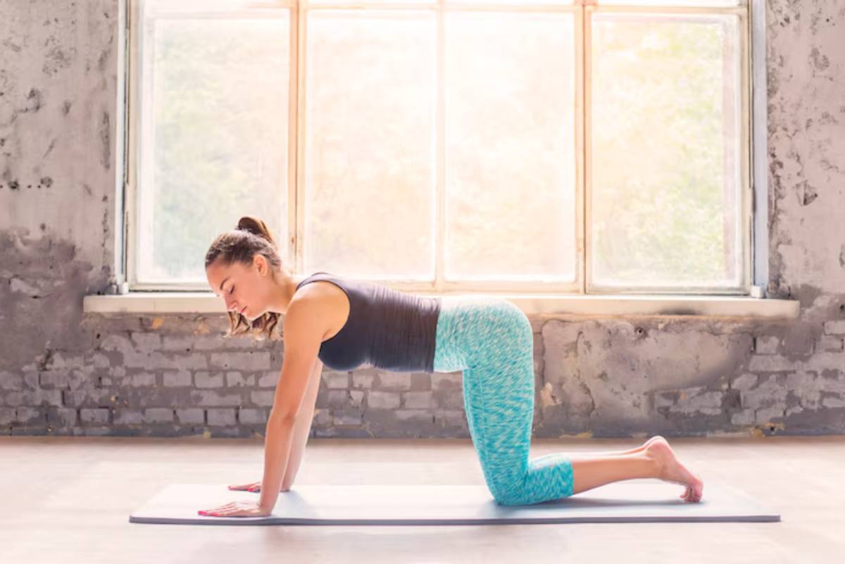 A person practices yoga on a mat indoors, in a bright room with large windows and a rustic wall backdrop.