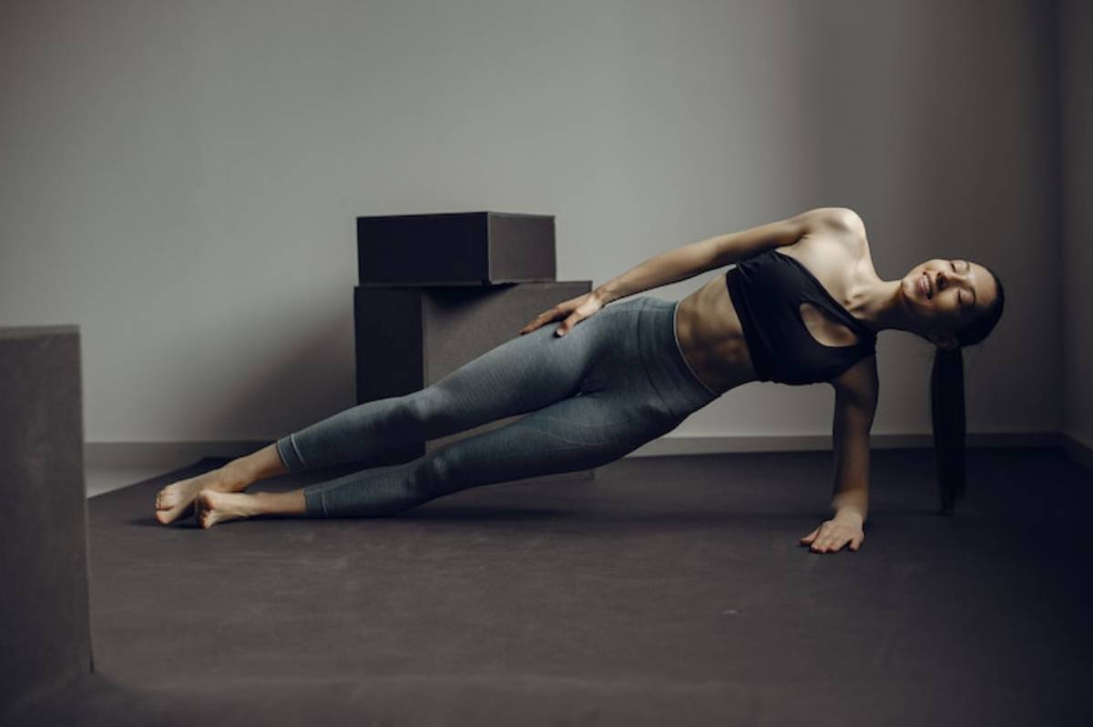 A person in a black crop top and gray leggings performs a side plank on a dark floor, surrounded by geometric shapes.