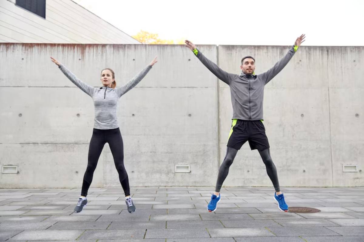 A man and woman energetically jump in unison, wearing athletic attire, against a concrete wall on a paved floor.