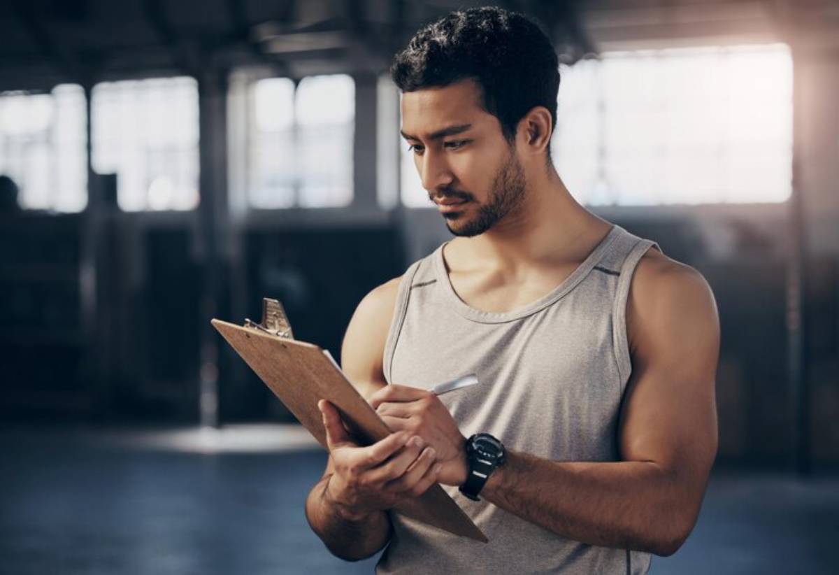 A muscular individual in a sleeveless shirt holds a clipboard and pen, focused in a well-lit gym environment.