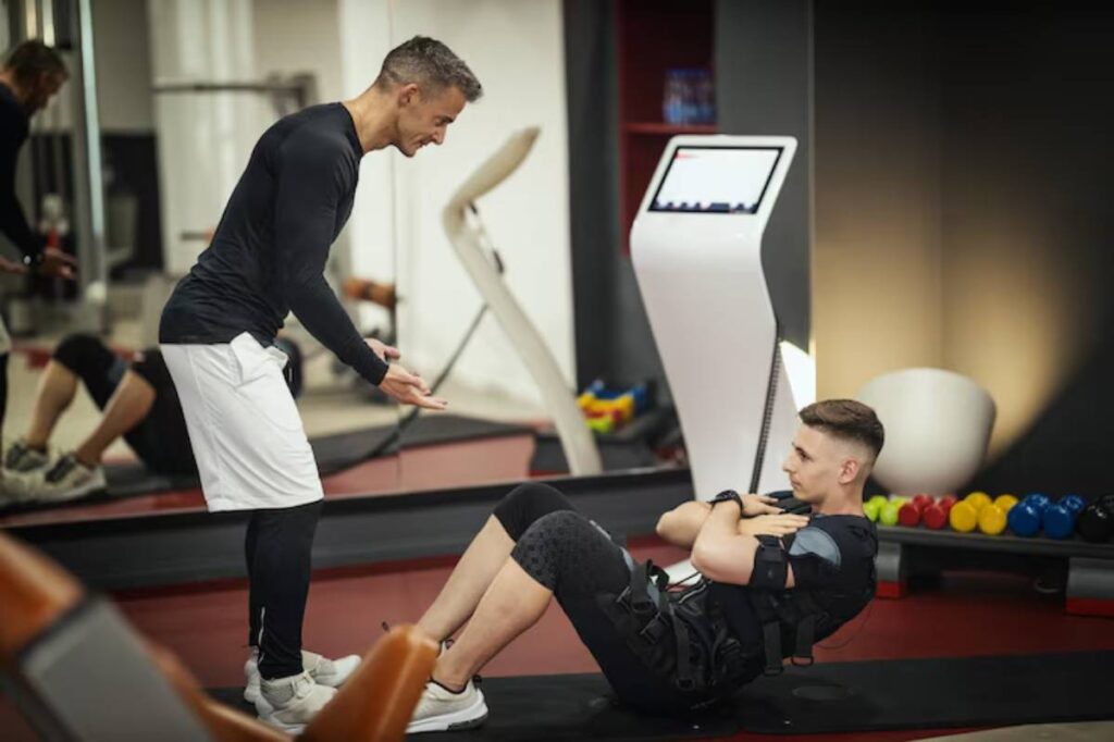 A trainer guides a client performing sit-ups on a mat in a modern gym, with exercise equipment and colorful weights in the background.