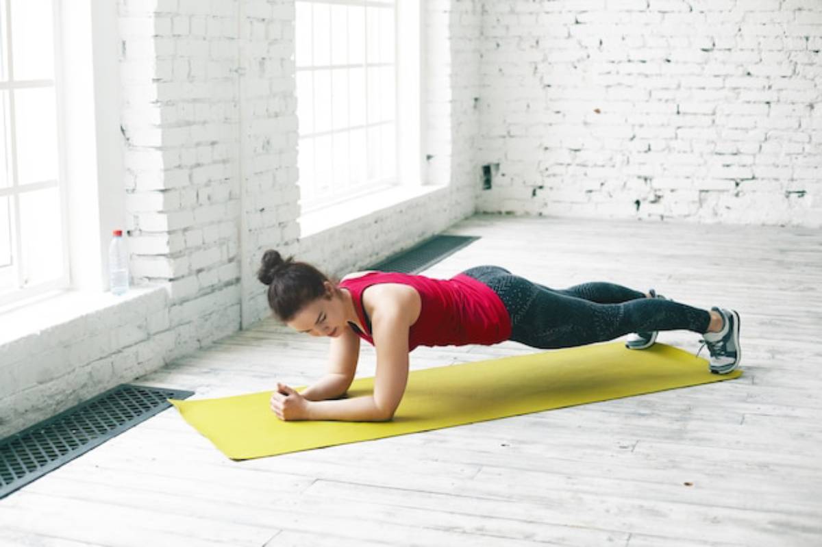 A person in a red tank top performs a plank exercise on a yellow mat in a bright, airy fitness space with white brick walls.