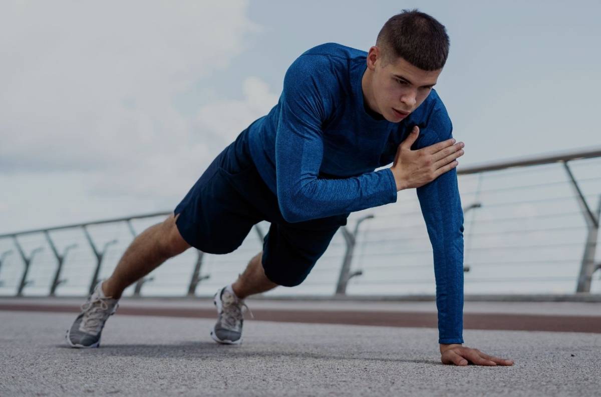 A man in a blue long-sleeve shirt and shorts performs a push-up on a pavement, with a railing and cloudy sky in the background.