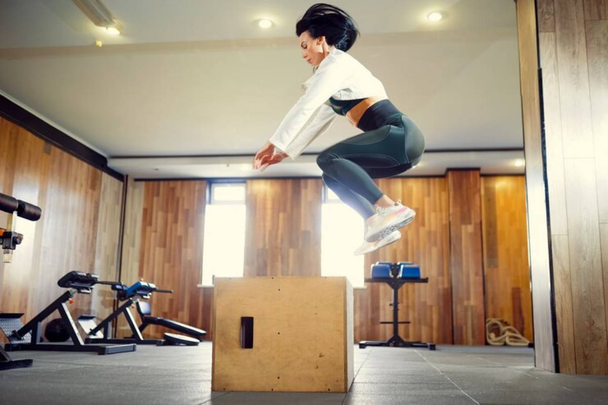 A person performs a box jump in a modern gym, surrounded by exercise equipment and natural light filtering through large windows.