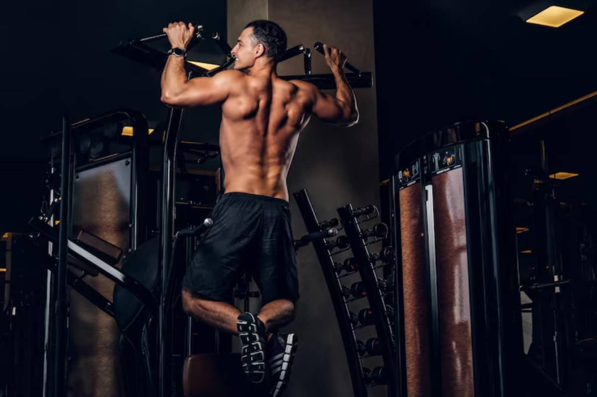 A muscular individual performs pull-ups at a gym, with fitness equipment visible in the background, highlighting strength training.