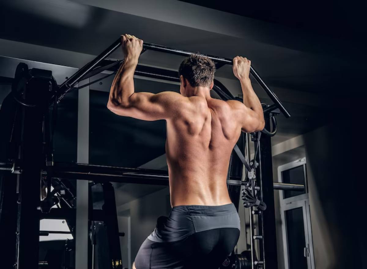 A muscular man performing pull-ups on a bar, showcasing his defined back and arms in a dimly lit gym.