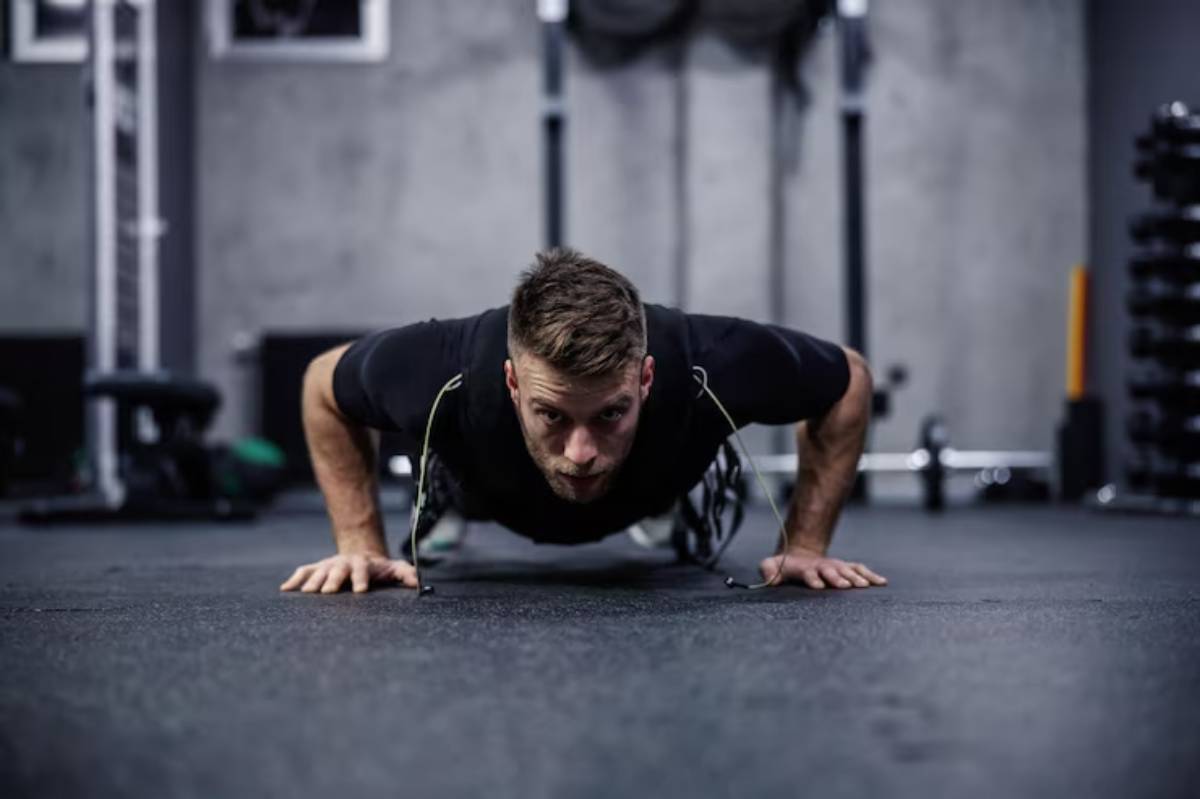 A person performs push-ups on a gym floor, surrounded by fitness equipment and weights in a modern workout space.