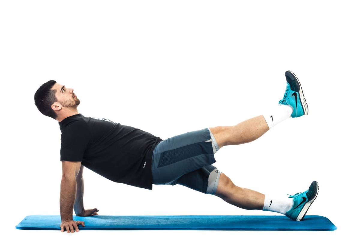 A person performs an exercise on a blue mat, balancing on one hand while extending a leg upwards, dressed in athletic wear.