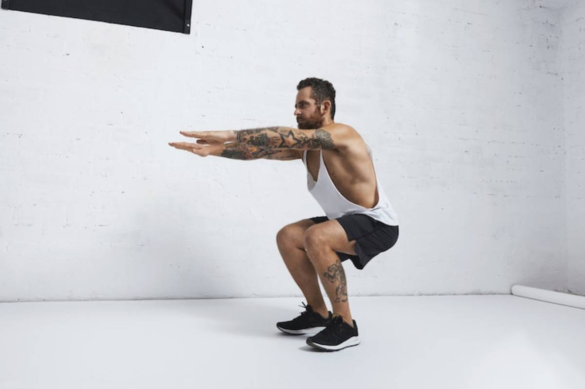 A muscular man in a tank top and shorts performs a squat exercise in a minimalist, well-lit gym setting.