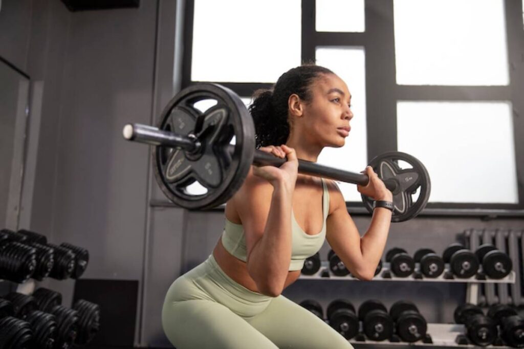 A woman in activewear performs a squat with a barbell in a modern gym, surrounded by dumbbells on shelves.