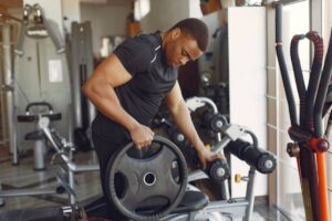 A person in a black athletic shirt adjusts a weight plate at a well-equipped gym with various workout machines in the background.