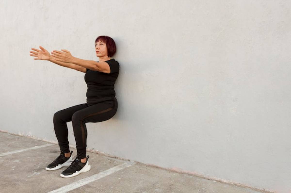 A person performs a wall squat against a plain wall, arms extended forward, dressed in black athletic wear and sneakers.