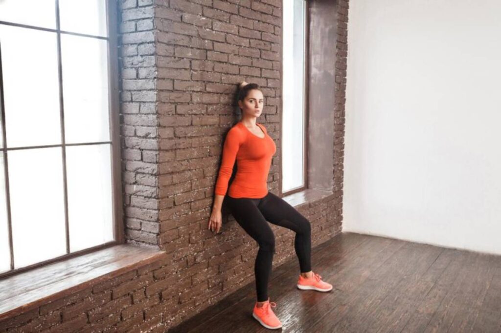 A woman in an orange long-sleeve shirt performs a wall sit exercise against a brick wall in a bright, minimalistic room.