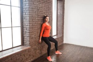 A woman in an orange long-sleeve shirt performs a wall sit exercise against a brick wall in a bright, minimalistic room.
