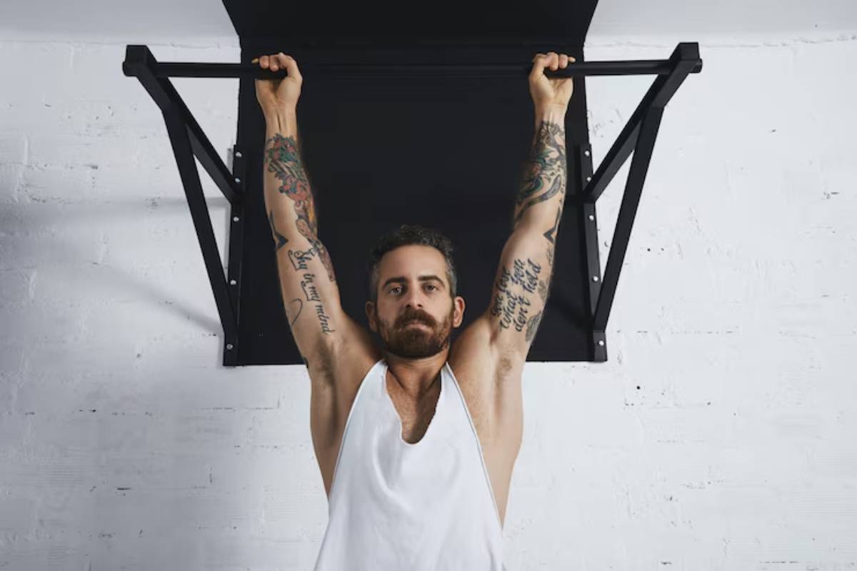 A muscular individual with extensive tattoos performs a pull-up on a black workout bar, set against a white brick wall.