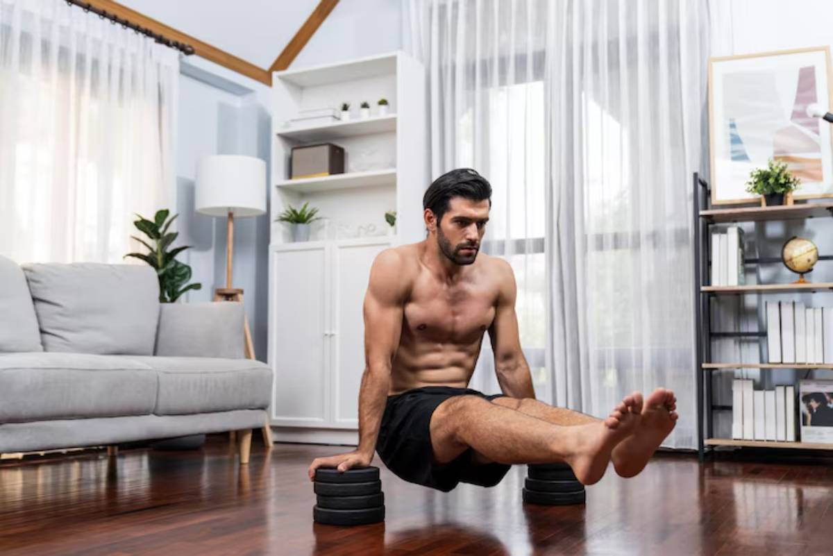 A muscular man performs an advanced exercise on wooden parallettes, balancing with legs extended and feet pointed, in a bright room.