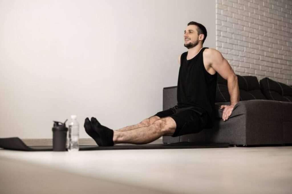 A person performing a workout, seated on a couch with legs extended, near a water bottle and fitness equipment on a mat.