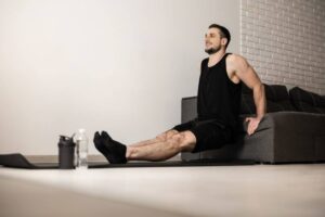 A person performing a workout, seated on a couch with legs extended, near a water bottle and fitness equipment on a mat.