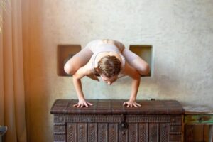 A person performs an intricate yoga pose on a decorative wooden chest, showcasing balance and flexibility in a serene indoor setting.