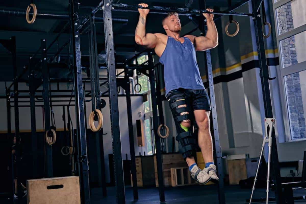 A muscular man performs a pull-up on gym rings, wearing a knee brace and shorts, with exercise equipment in a dimly lit gym.