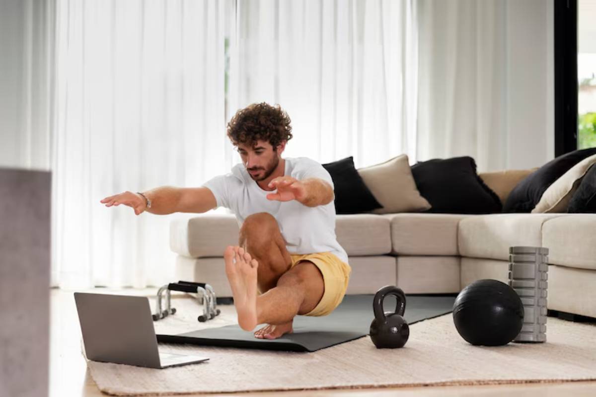 A person performs an exercise on a mat at home, with a laptop in front and various fitness equipment nearby. Bright, airy space.