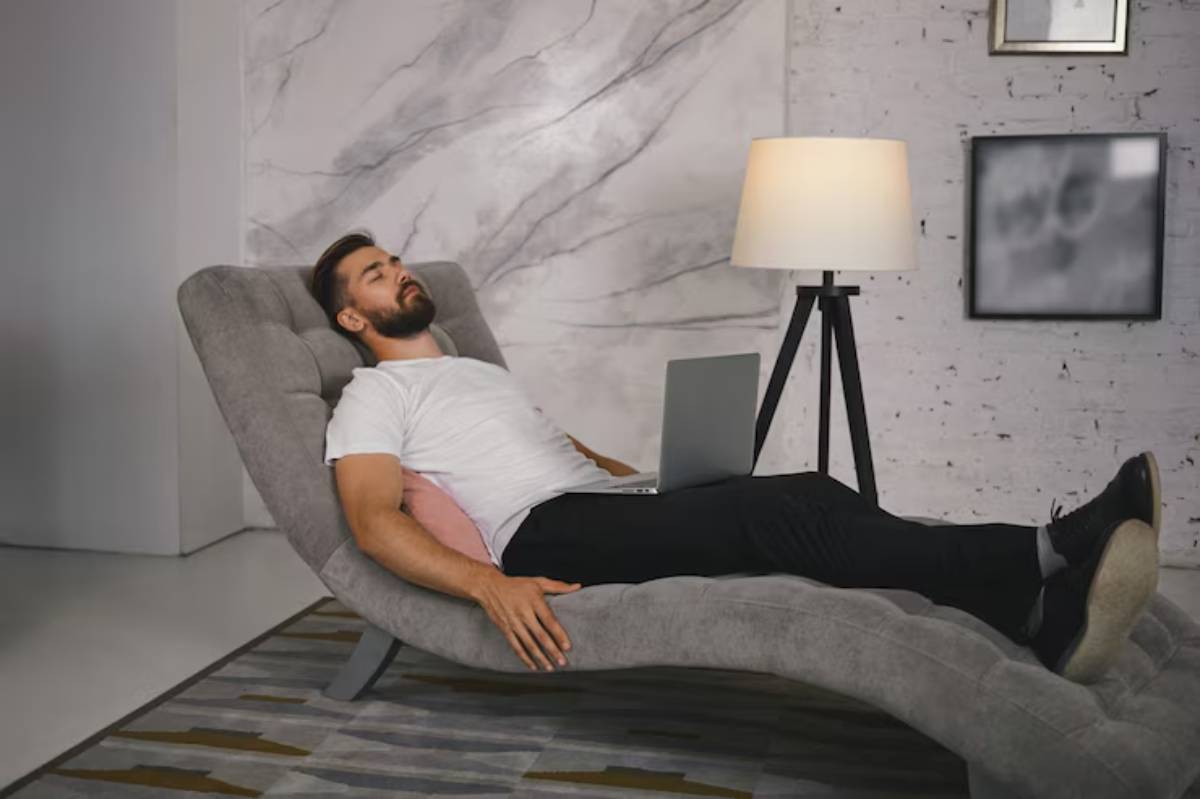 A person relaxes on a modern, curved gray lounge chair, working on a laptop with a floor lamp and abstract wall art in the background.
