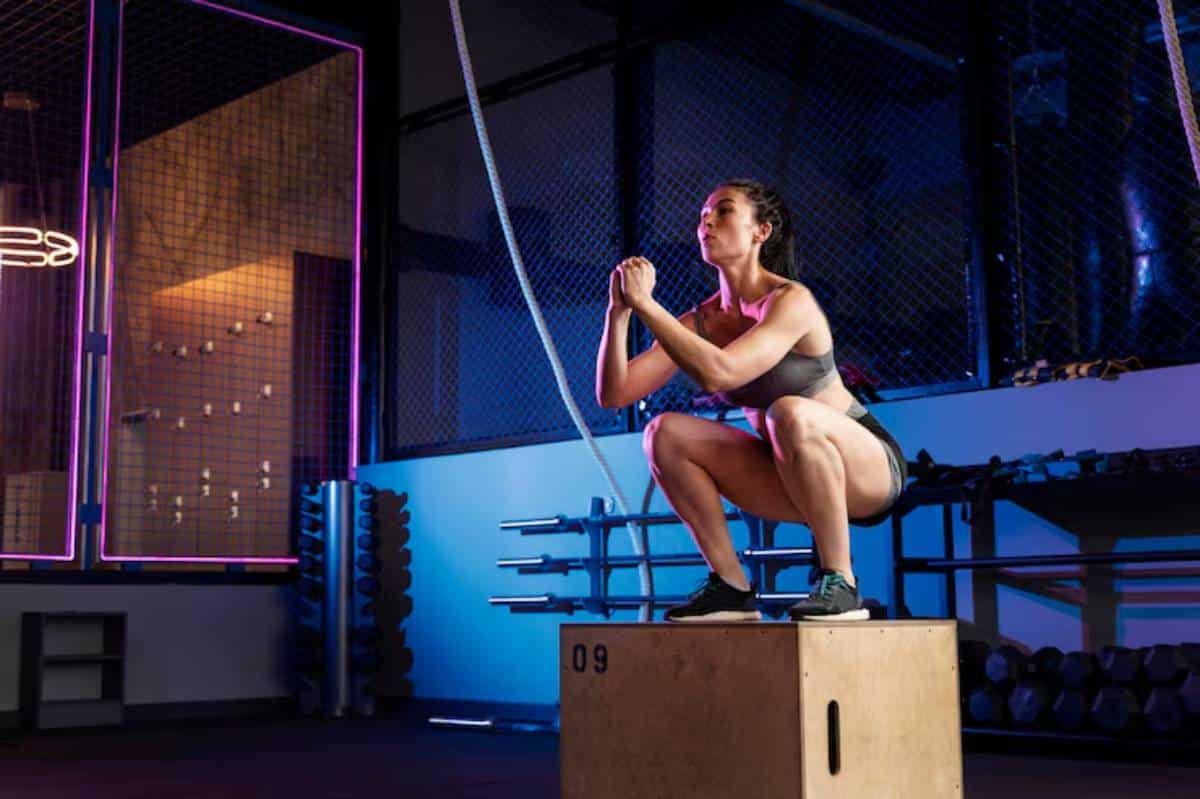 A person performs a squat on a wooden box in a vibrant gym with blue and purple lighting, surrounded by fitness equipment.