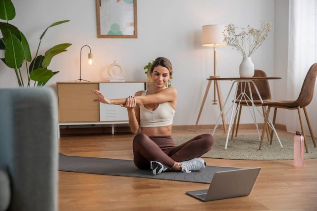 A person in a yoga outfit sits on a mat, stretching their arm in a well-lit, modern living room with plants and decor.