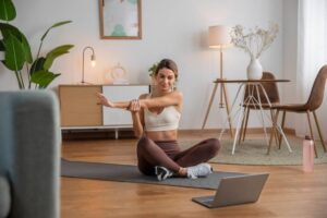 A person in a yoga outfit sits on a mat, stretching their arm in a well-lit, modern living room with plants and decor.