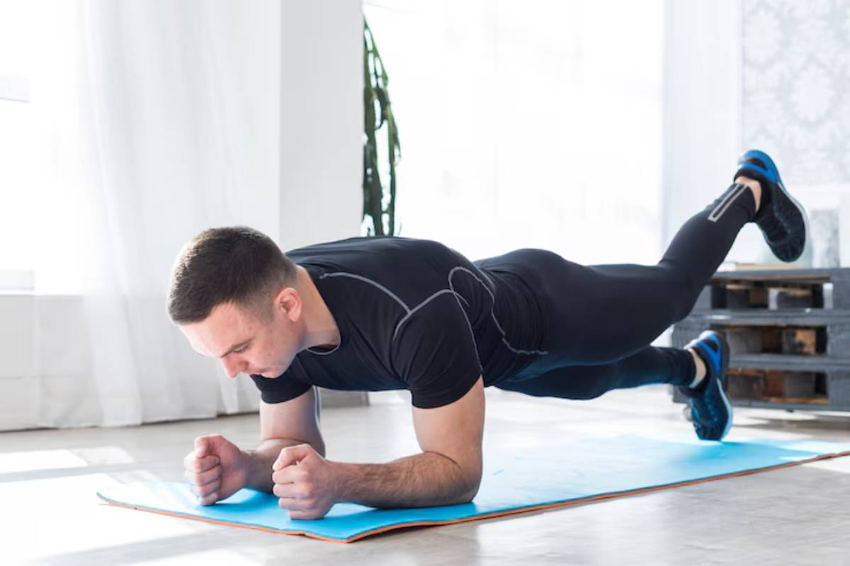 A man in athletic wear performs a plank exercise on a blue mat in a bright, airy room with large windows and greenery.