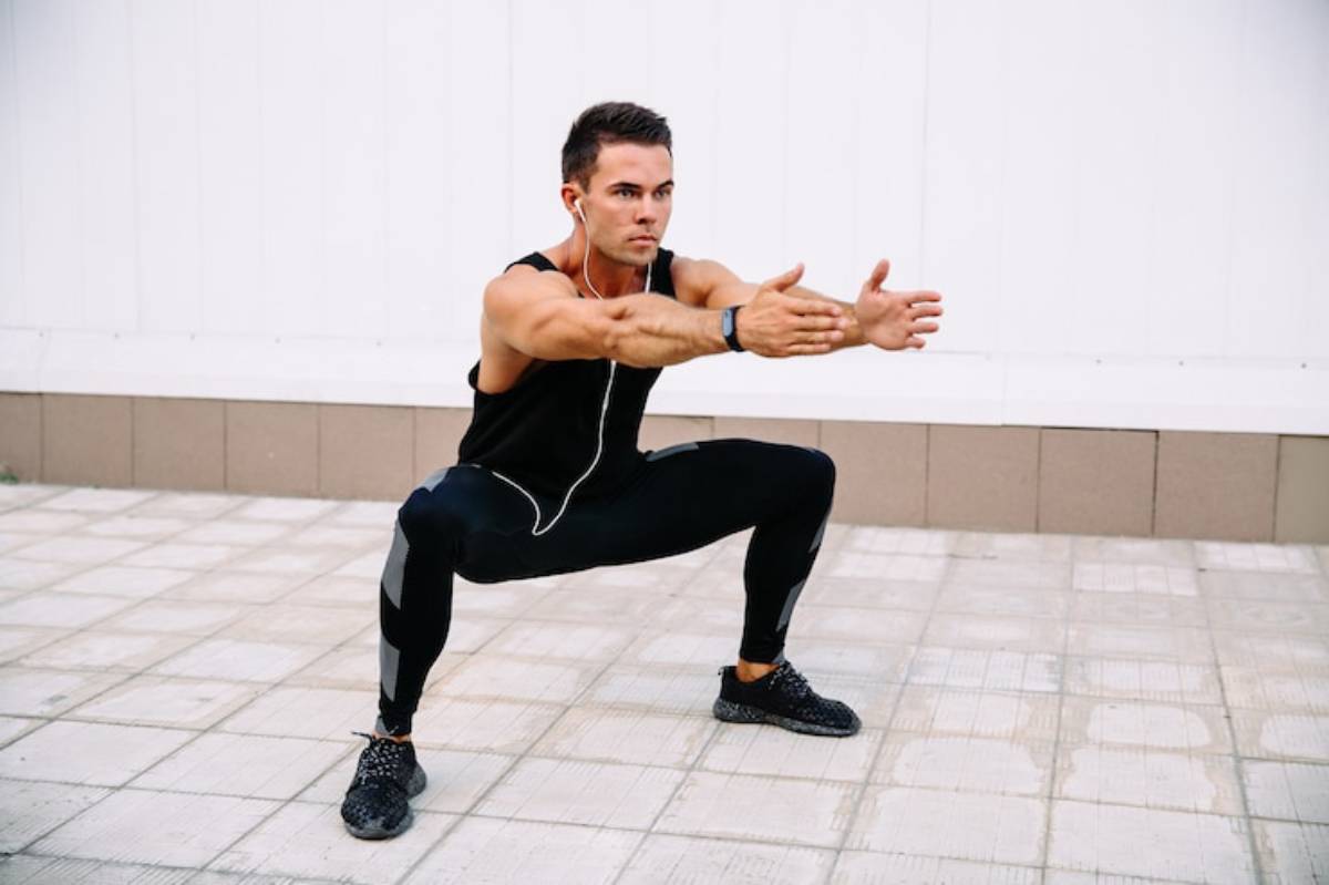 A person performs a squat exercise outdoors, wearing workout clothes and earbuds, with arms extended forward.
