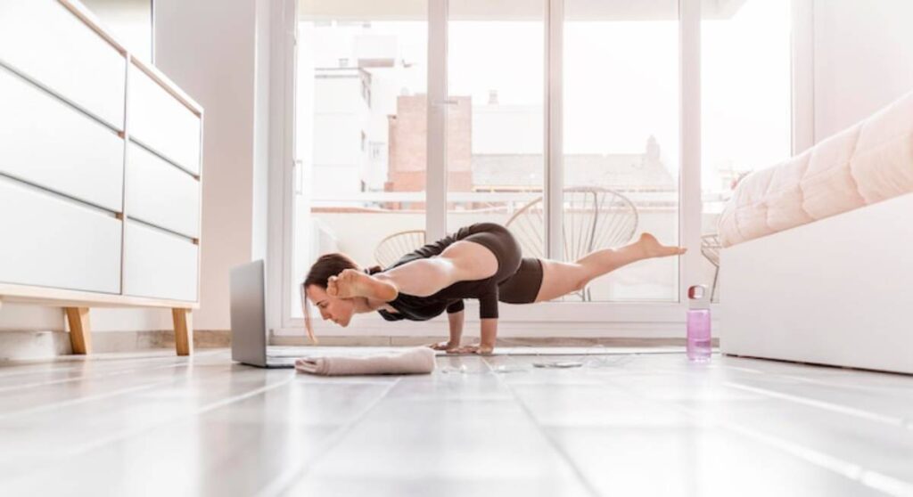 A person practicing yoga in a bright room, balancing in a pose with a laptop in front, towel and water bottle nearby.