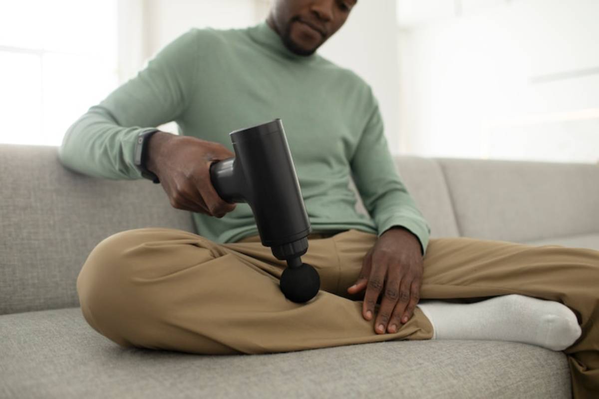 A person in a green sweater sits cross-legged on a couch, using a black massage gun on their thigh for muscle relief.