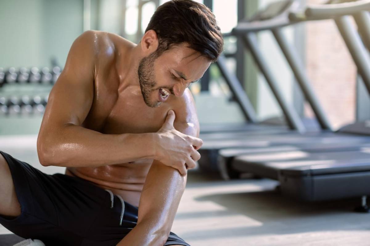 A shirtless man in a gym sits on the floor, holding his shoulder with a concerned expression, surrounded by fitness equipment.