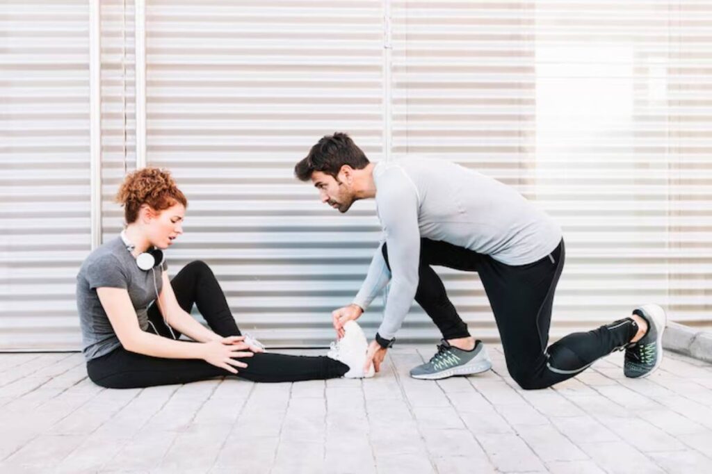 A man in athletic wear assists a seated woman in stretching her leg, both appearing focused in a bright, modern space.