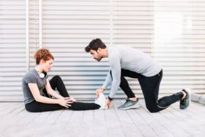 A man in athletic wear assists a seated woman in stretching her leg, both appearing focused in a bright, modern space.
