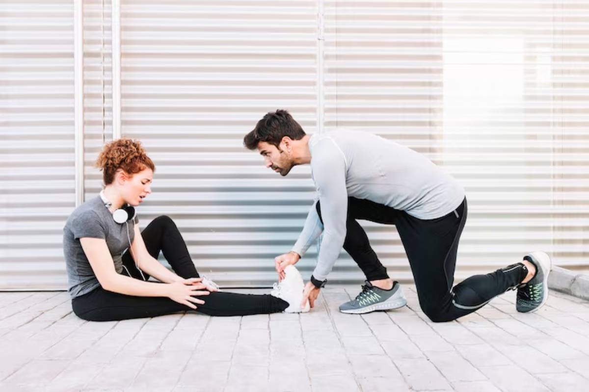A man in athletic wear assists a seated woman in stretching her leg, both appearing focused in a bright, modern space.