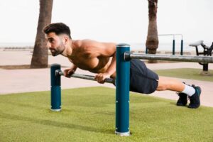 A muscular individual performs a push-up on parallel bars, set on a grassy area in an outdoor fitness space with palm trees in the background.