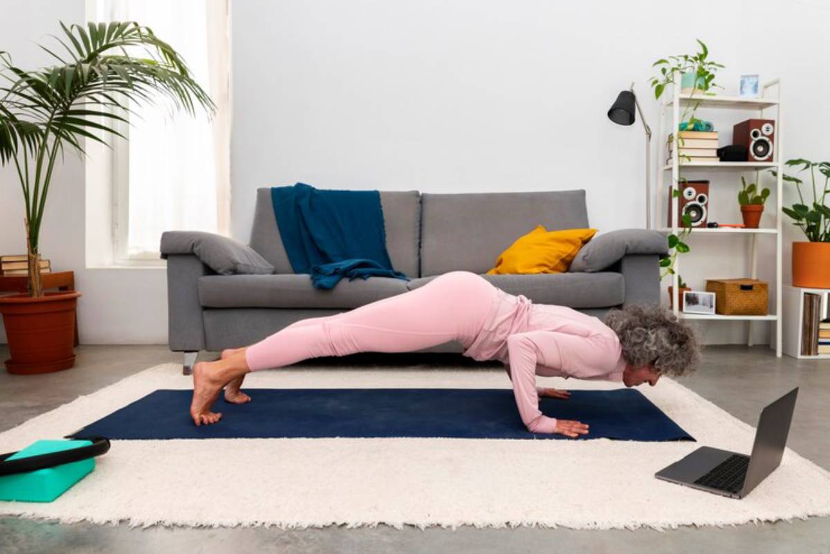 An older woman in pink workout clothes performs a yoga pose on a mat at home, with a laptop nearby.