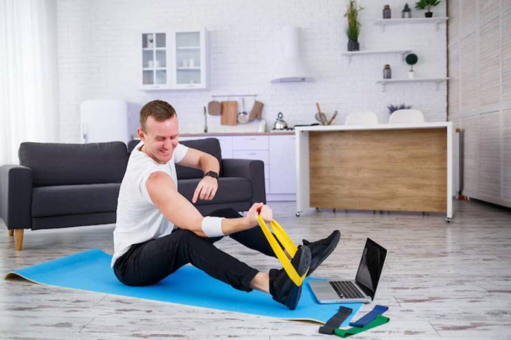 A person exercises on a blue mat at home, using a resistance band while a laptop and various bands are nearby.
