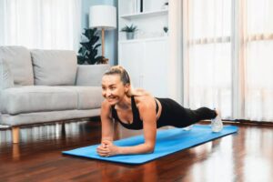 A person performs a plank exercise on a blue mat in a bright living room with a gray couch and potted plants.