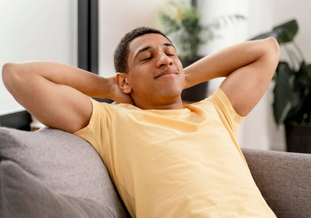 A person relaxes on a gray couch, wearing a yellow shirt, with arms behind their head, in a modern, bright indoor space.