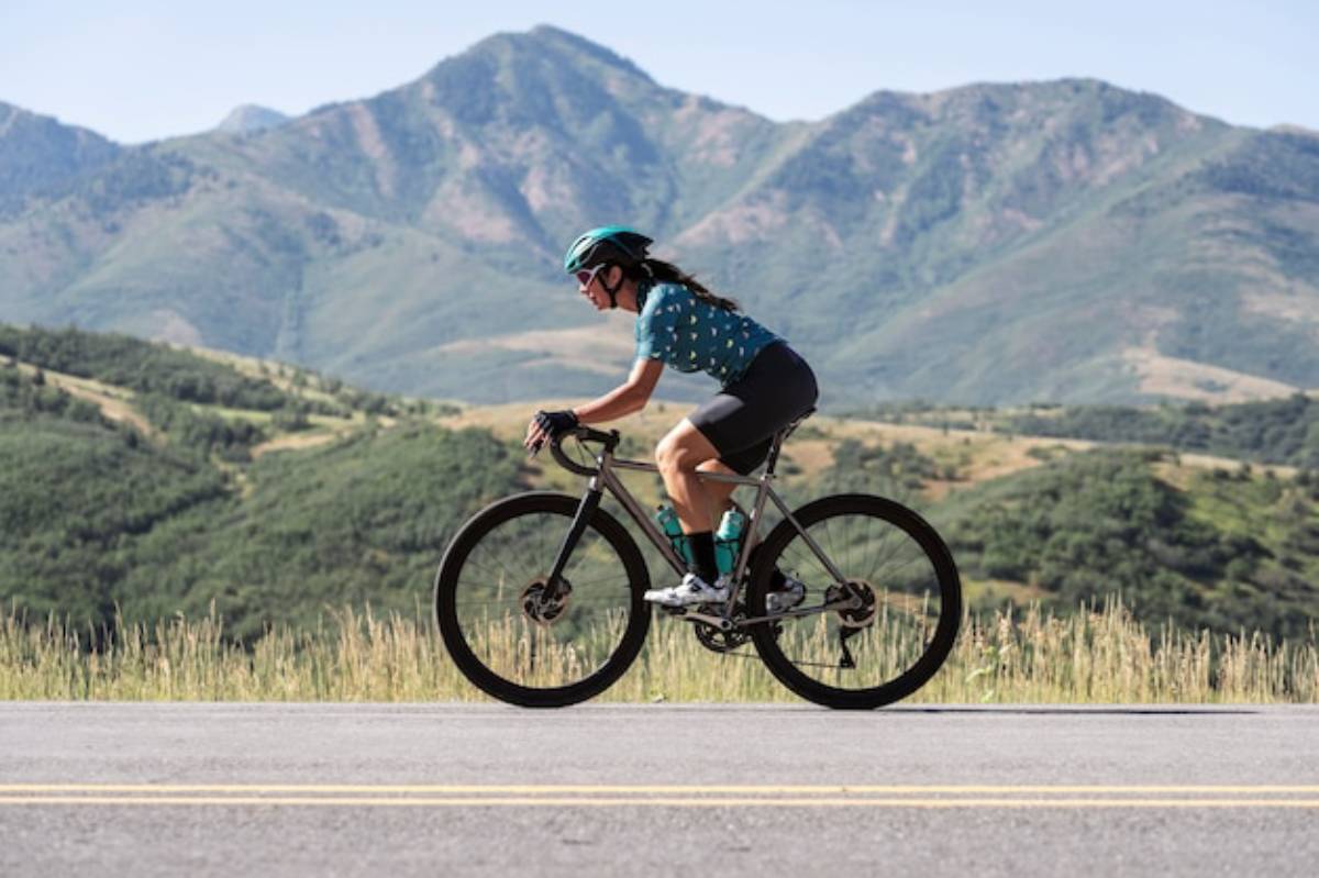 A cyclist rides on a road with green hills and mountains in the background, enjoying a sunny day outdoors.