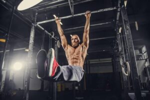 A muscular individual performs a pull-up on a gym rig, showcasing strength against a backdrop of workout equipment and soft lighting.